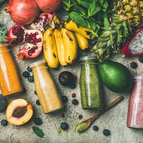 Flat-lay of colorful smoothies in bottles with fresh tropical fruit and vegetables on concrete background, top view, square crop. Healthy, vegetarian, detox, dieting breakfast food concept